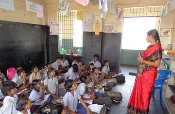 A standing woman in a red sari teaches rows of young uniformed children seated on the floor in a primary school classroom in India.