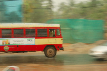 A panning shot of a KSRTC bus moving from left to right. A smaller car, blurred, is seen coming from the right to left, veering away from the bus. It is a rainy day, and some construction site can be seen on the other side, behind the two vehicles.