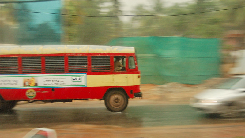 A panning shot of a KSRTC bus moving from left to right. A smaller car, blurred, is seen coming from the right to left, veering away from the bus. It is a rainy day, and some construction site can be seen on the other side, behind the two vehicles.