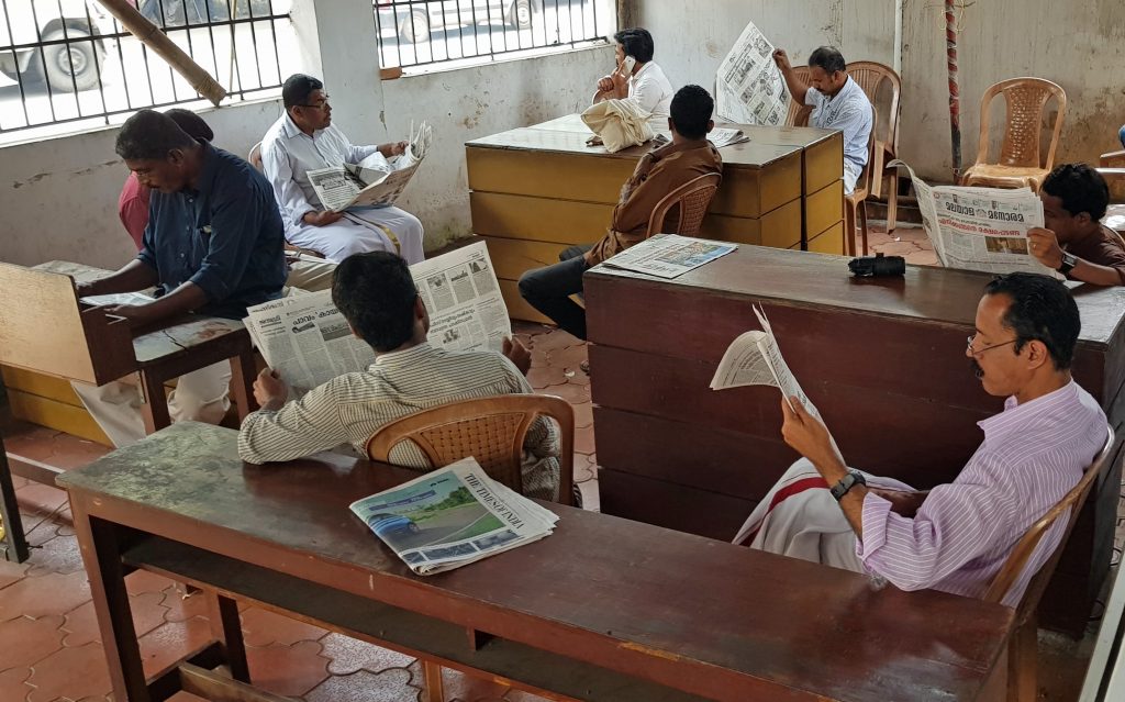 A photograph of a reading room in Kerala, where seven men sit around at benches, reading newspapers.
