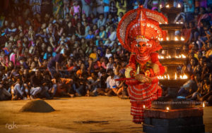 An image of the theyyam performance. A large crowd is seen off focus, in the background. In focus, towards the foreground, is the theyyam, left of frame. Also in focus, in front of the theyyam is a large lit lamp. 