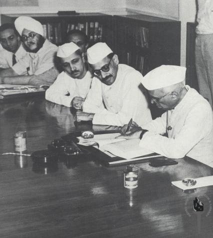 Nehru signing the FYP (right), as five others look on, seated to his right in the image. K.N. Raj seen at the far end of the table.