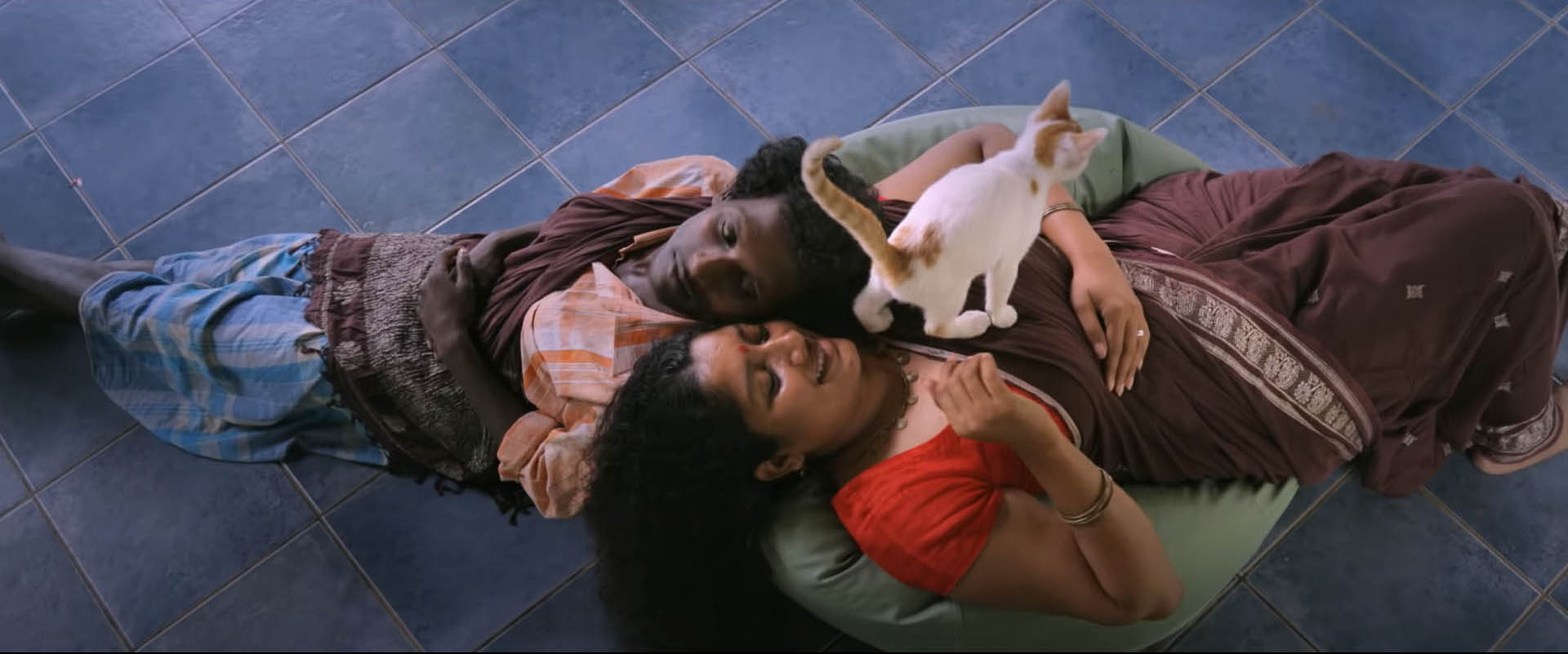 Top-angle shot of Gulikan and Dancer--two characters form the film--lying on a beanbag with their heads next to each other, stretched out in opposite directions, with a cat standing on the Dancer's chest
