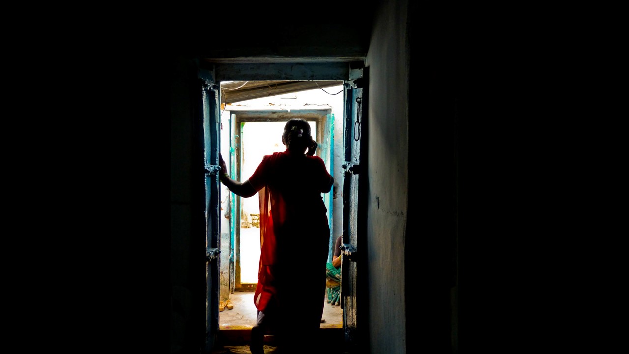 A silhouette of a woman standing near a door, her hands resting on the frame of the door to her left; she faces away from the camera