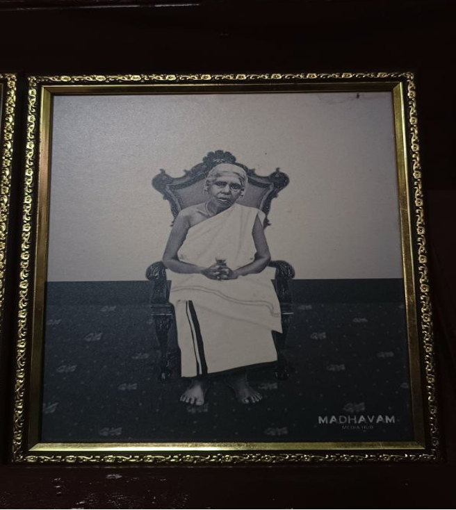 A photograph of a gold-framed photo, in balck and white, of an old woman in a white set mundu, seated on an ornate chair.
