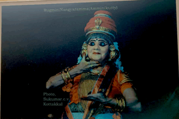 A photograph of an older woman in Nangiarkoothu costume and makeup, mid-pose onstage.
