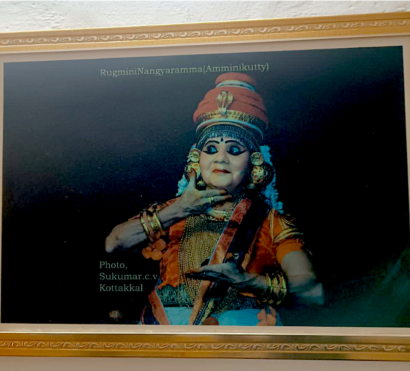 A photograph of an older woman in Nangiarkoothu costume and makeup, mid-pose onstage.